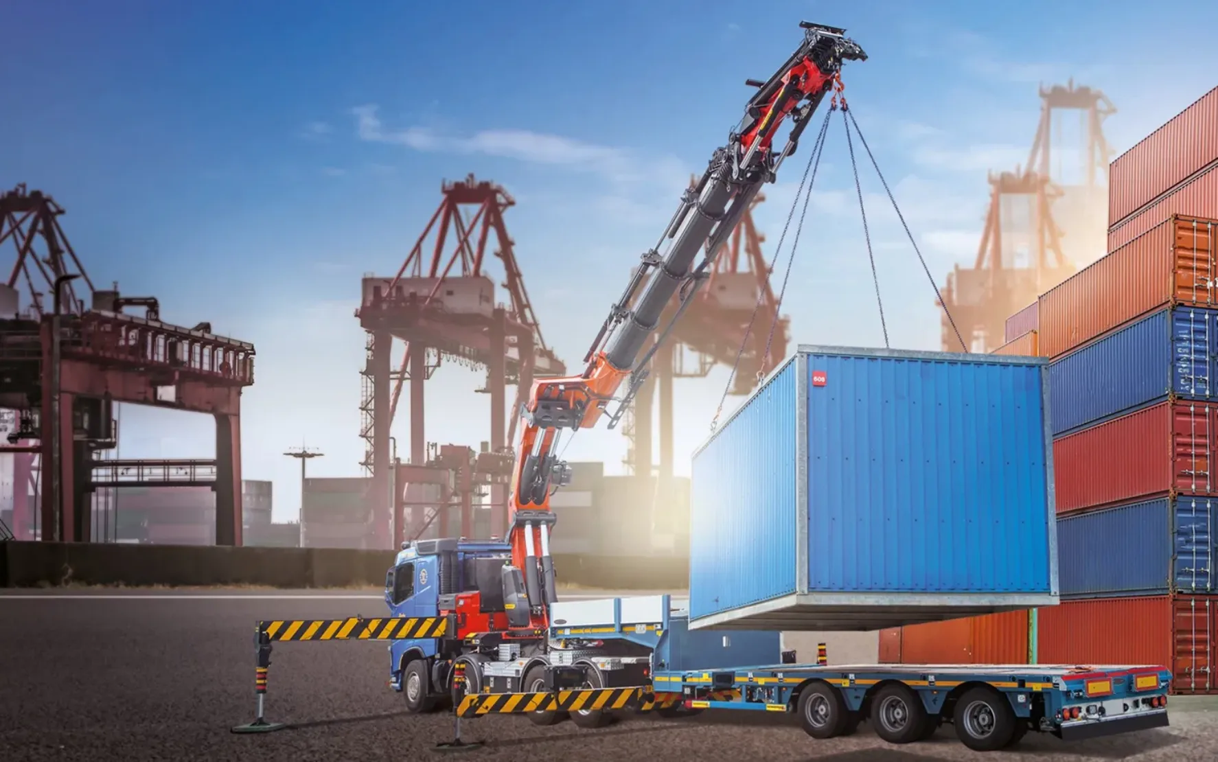 headerimage Ein Kranwagen hebt in einem großen Verladehof einen blauen Frachtcontainer auf einen Anhänger. Im Hintergrund sind Stapel bunter Container und Frachtkräne unter einem klaren Himmel zu sehen.